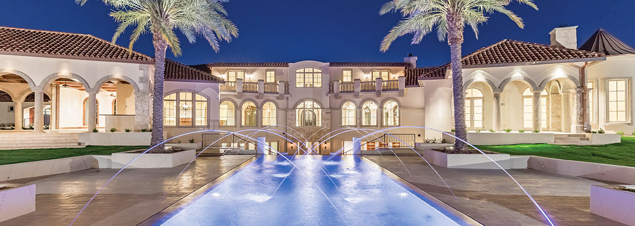 Lit Mediterranean-style desert mansion behind a fountain courtyard at dusk