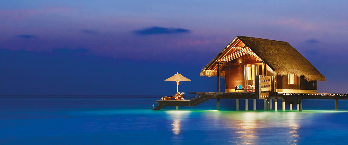 Thatched overwater bungalow lit at dusk above calm turquoise water