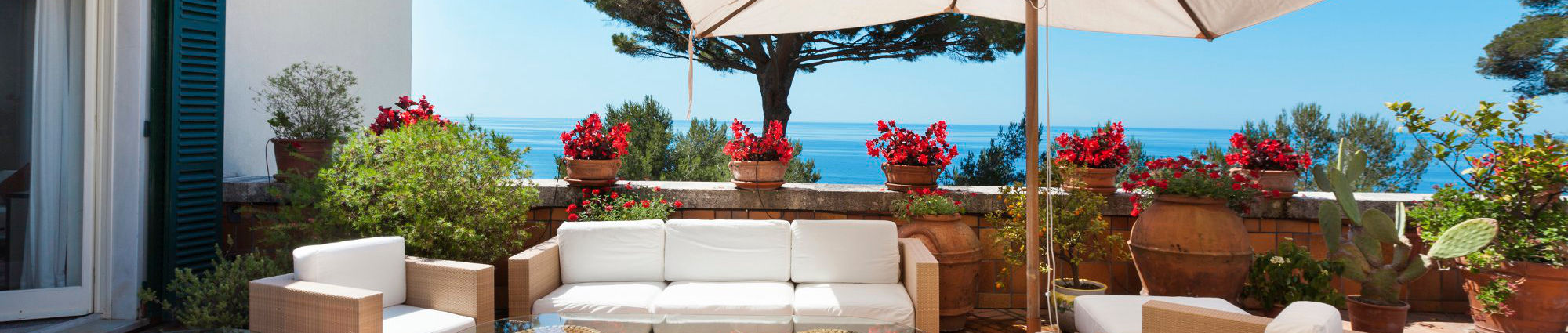 Mediterranean terrace with potted geraniums and an umbrella looking out to a blue sea