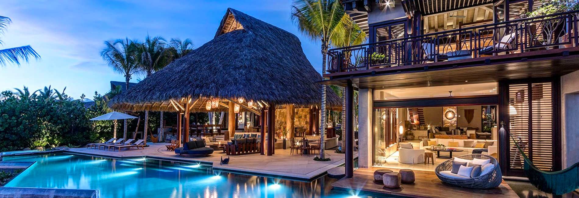 Thatched-roof villa beside a lit swimming pool among palms at dusk
